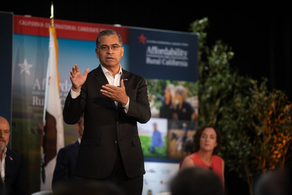 A person in a black suit gestures with their hands as they stand on a stage in front an audience. Other panelists can be seen in the background, along with a banner that reads “Affordability and Rural California.”