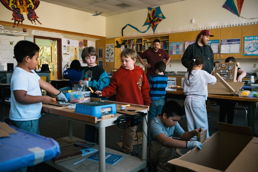 Young children work with various school supplies inside a classroom as two instructors stand nearby.