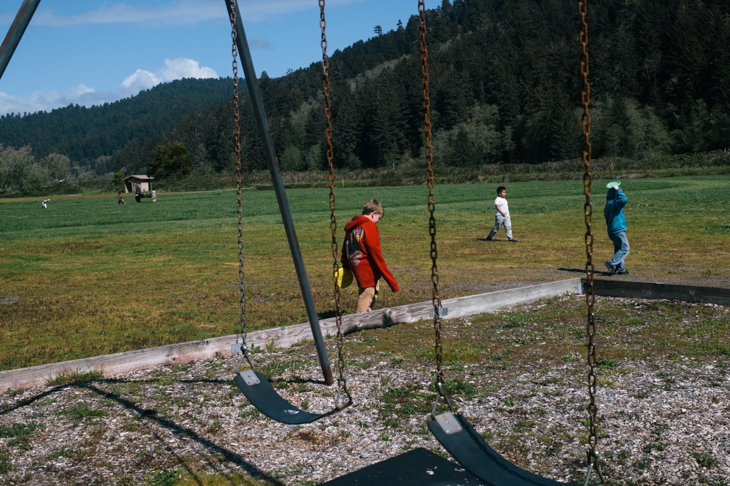 Three students play frisbee on an open grass field overlooking mountain ranges filled with pine trees. A swig set can be seen in the foreground.