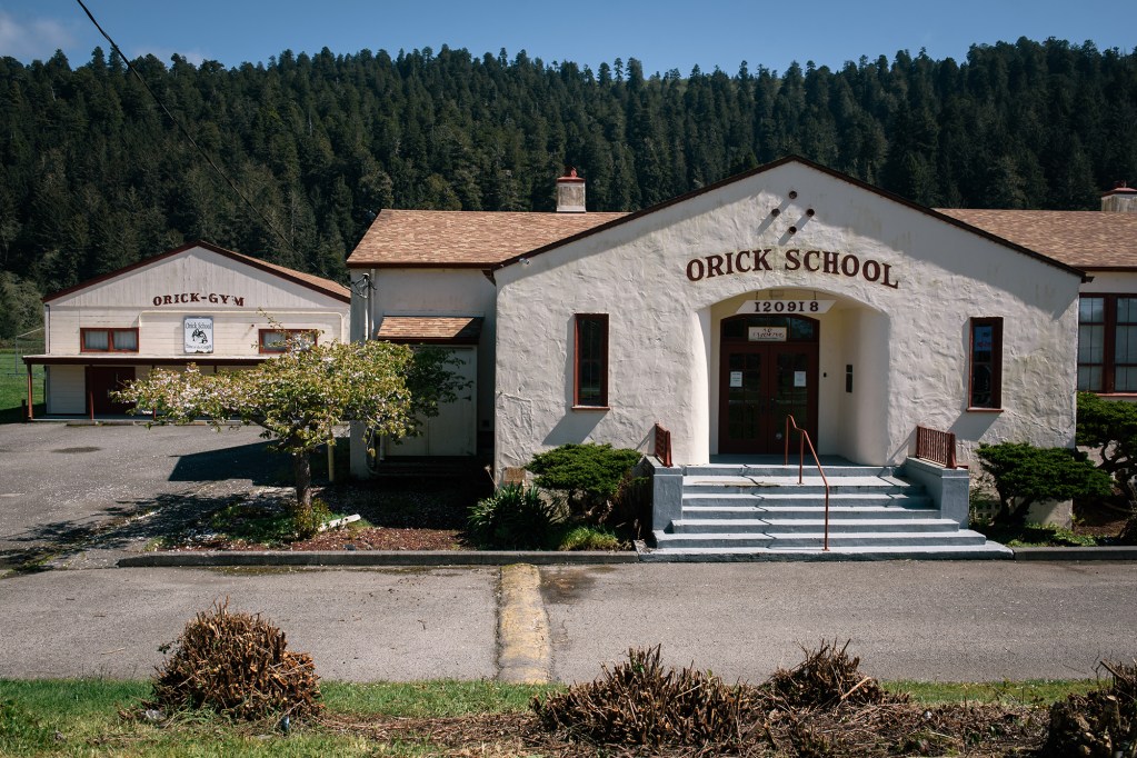 A beige school building with red accent details overlooking a small mountain range filled with pine trees. The school building includes a label that reads "Orick School."