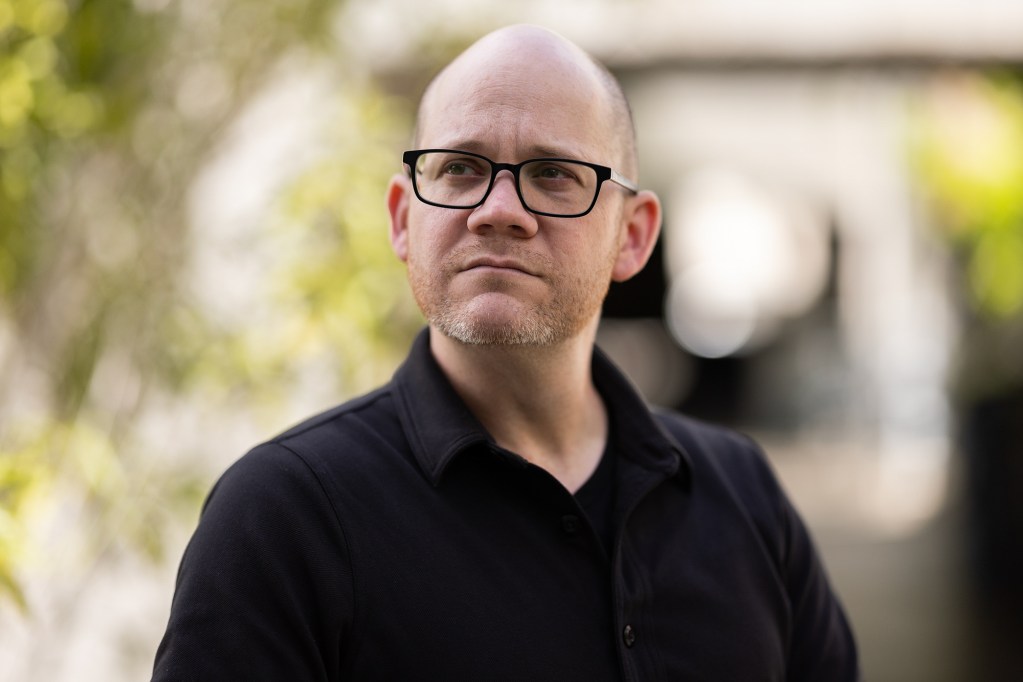 A person wearing black glasses and shirt looks off into the distance while standing on a college campus.