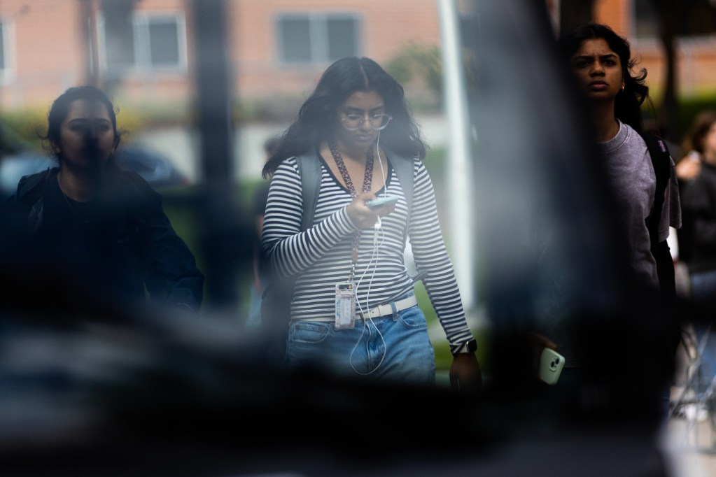Three students walk through a campus. The photo focus on the student in the middle, who wears a black and white striped shirt and looks down at their phone.