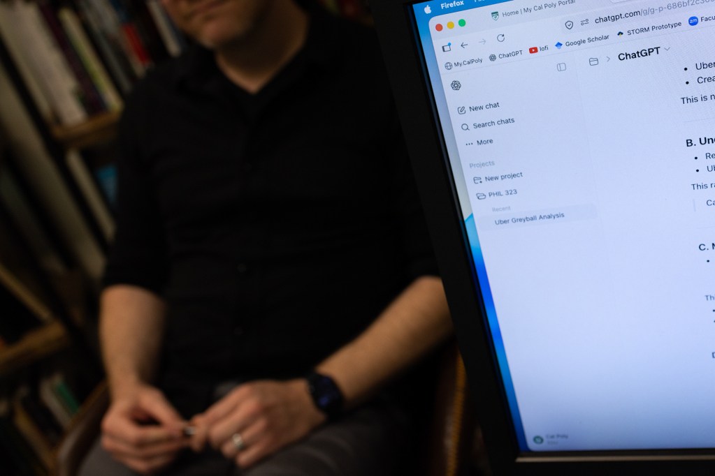The edge of a laptop that displays the ChatGPT website, as a person sits on a chair in the background.