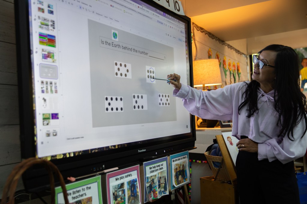 An over-the-shoulder view of a person in a white button-up shirt in a brightly colored, decorated classroom, with a large television screen displaying a math problem.