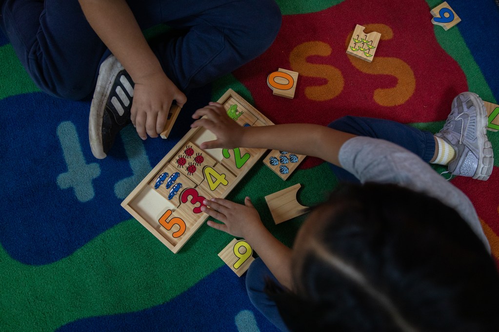 A top-down view of a pair of children's hands playing with a set of blocks with numbers on them in a carpeted area of a classroom,