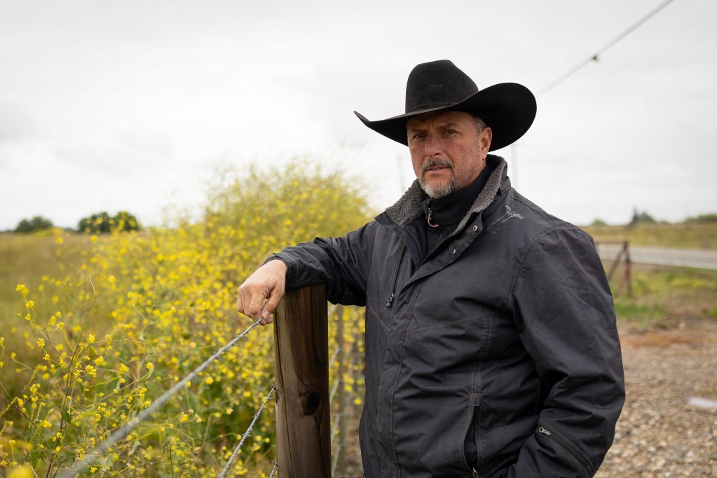 A person wearing a black cowboy hat and jacket stands beside a wooden fence post and barbed wire, with yellow wildflowers and open land in the background.