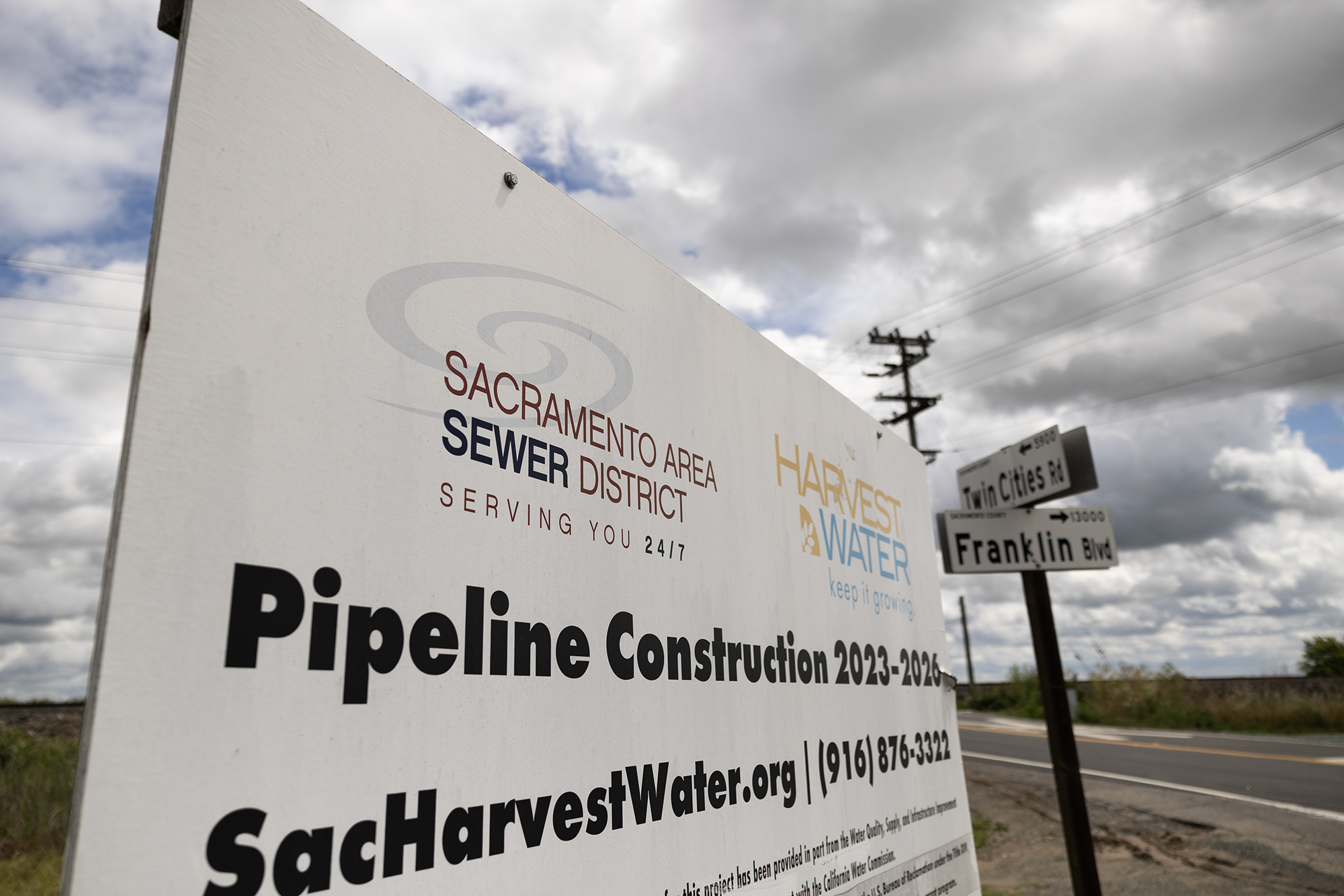 A roadside sign for the Sacramento Area Sewer District announces “Pipeline Construction 2023–2026,” with nearby street signs for Twin Cities Road and Franklin Boulevard under a cloudy sky.