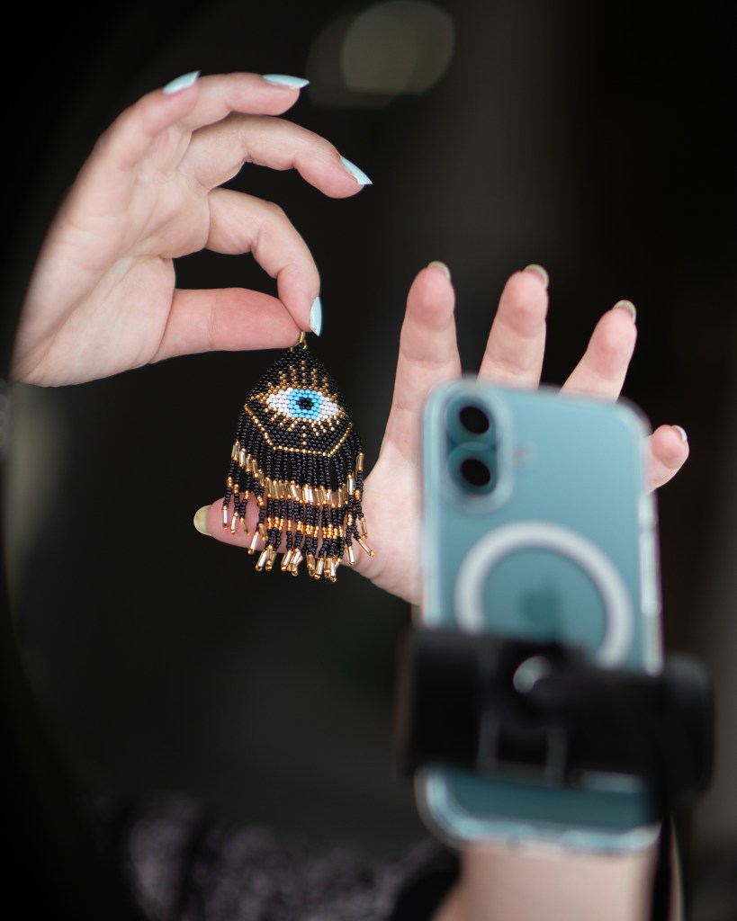 A close-up view of a person's hand dangling a black and blue eye-shaped beaded earring in front of ring lights and a cell phone.