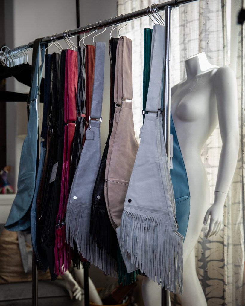 Several colorful cross-body leather bags hang on on a clothing rack near an undressed mannequin.