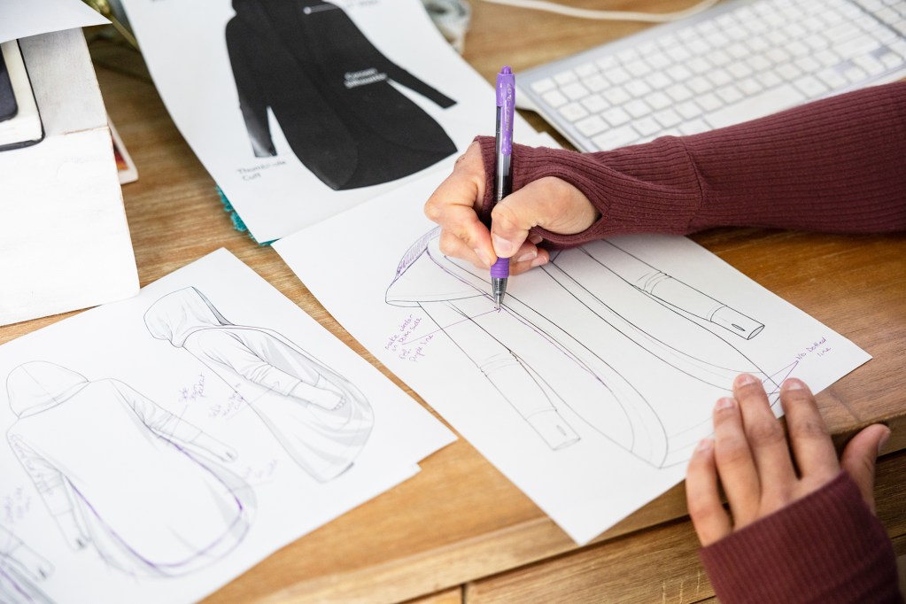 A close-up of a person's hand as they hold a purple pen and write on a sketch of a jacket design that lays on a wooden desk.