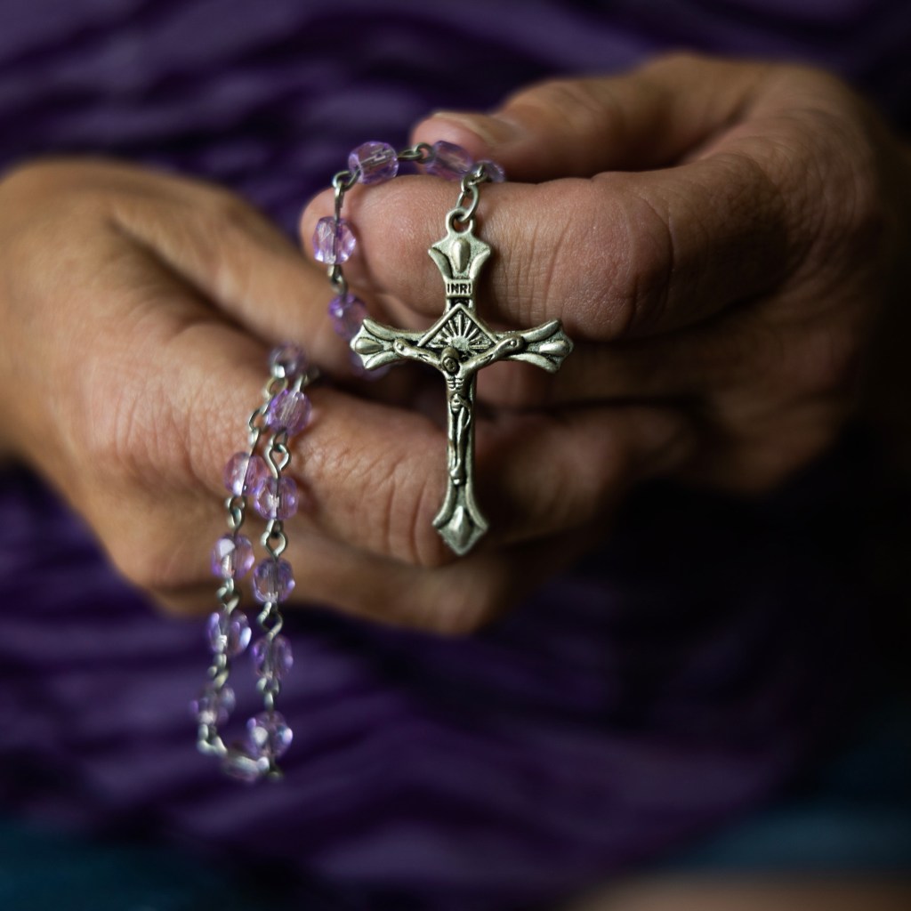 Hands hold a rosary with purple beads and a small metal crucifix.