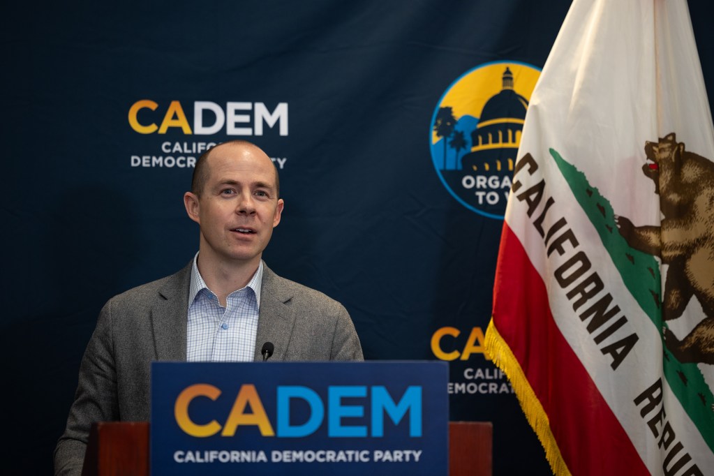 A person, wearing a gray blazer with a flannel shirt, stands in front of a podium as they speak to members of the media. A blue step-and-repeat with the California Democratic Party log can be seen behind the person, next to the California flag.
