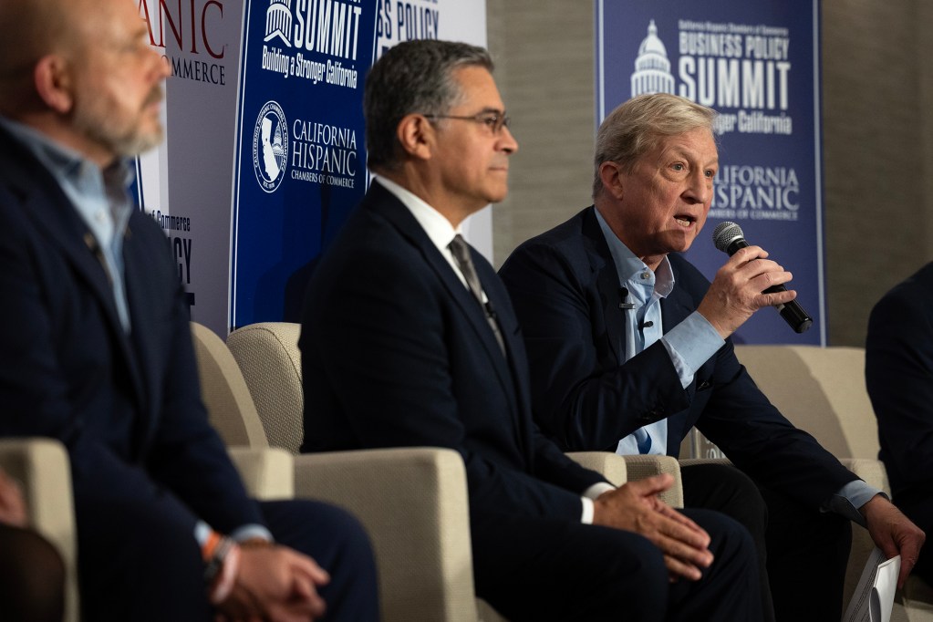 Several people sit onstage during a policy forum in front of signs for the California Hispanic Chambers of Commerce Business Policy Summit. One speaker holds a microphone while speaking as the others listen from nearby chairs.