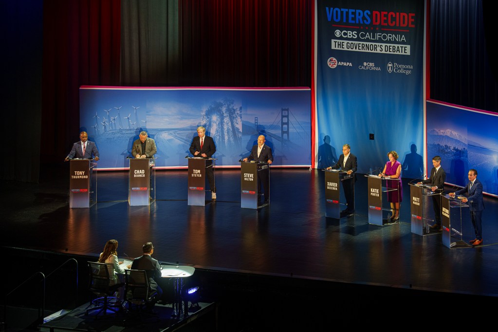 Eight candidates stand at separate podiums across a stage during a televised California governor’s debate, with moderators seated at a table in the foreground and a large “Voters Decide” backdrop behind them.