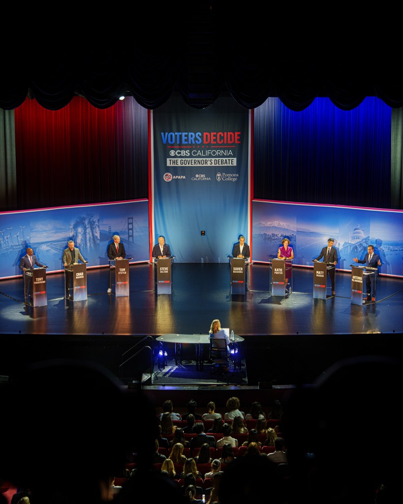 Eight candidates stand at separate podiums across a stage during a televised California governor’s debate, with moderators seated at a table in the foreground and a large “Voters Decide” backdrop behind them.