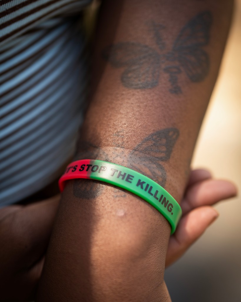 A close-up shows a person’s forearm with butterfly tattoos and a red-and-green silicone bracelet that reads “STOP THE KILLING.” The person’s hands rest together in sunlight, with part of a striped shirt visible at the edge of the frame.