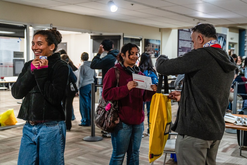 A person wearing a brown sweatshirt and a pair of headphones around their neck is placing a ballot into a yellow protective bag held by a staffer at a voting center. In the background, a line of people waits to fill out their ballots at the voting center.