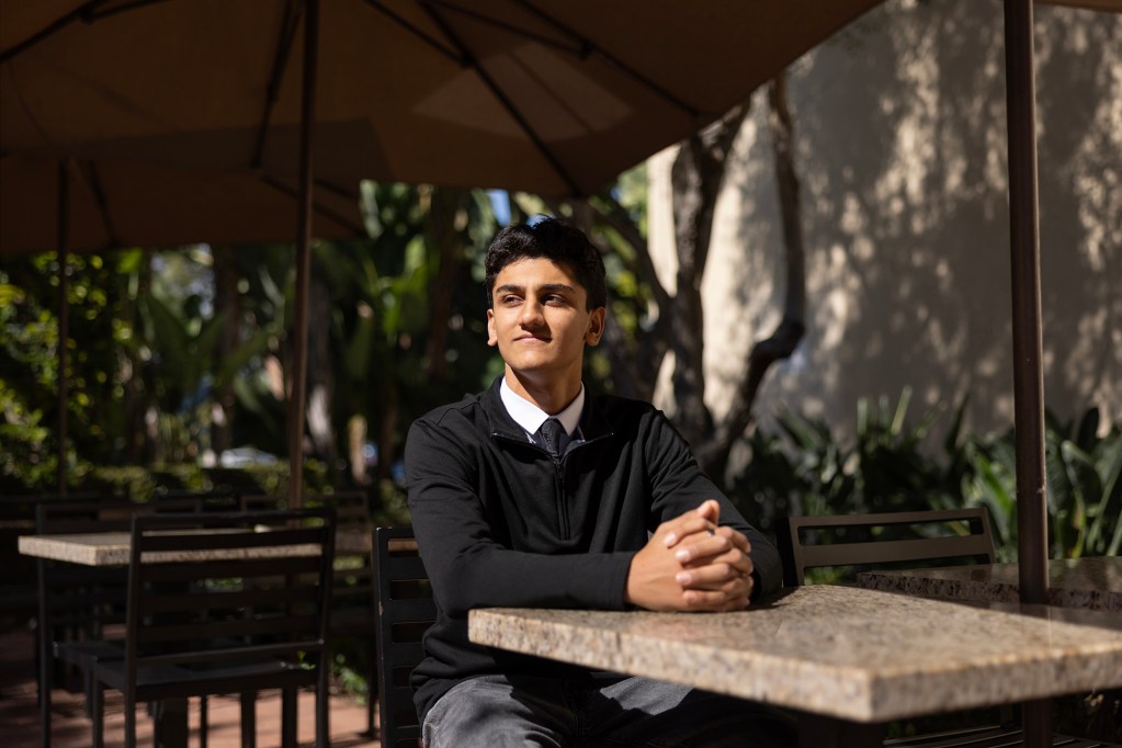 A person wearing a dark sweater with a dress-up shirt and tie underneath sits at a table in a patio area, their hands cupped together, looking away.