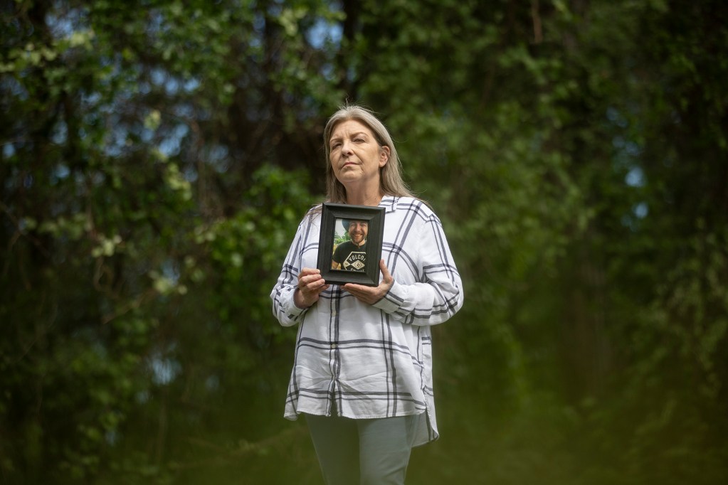 A person with grey hair wearing a white flannel shirt holds up a wooden framed photo of a person to their chest with both hands, In the background is a wall of green foliage from a row of trees behind the subject with specks of blue sky peaking though breaks in the branches.