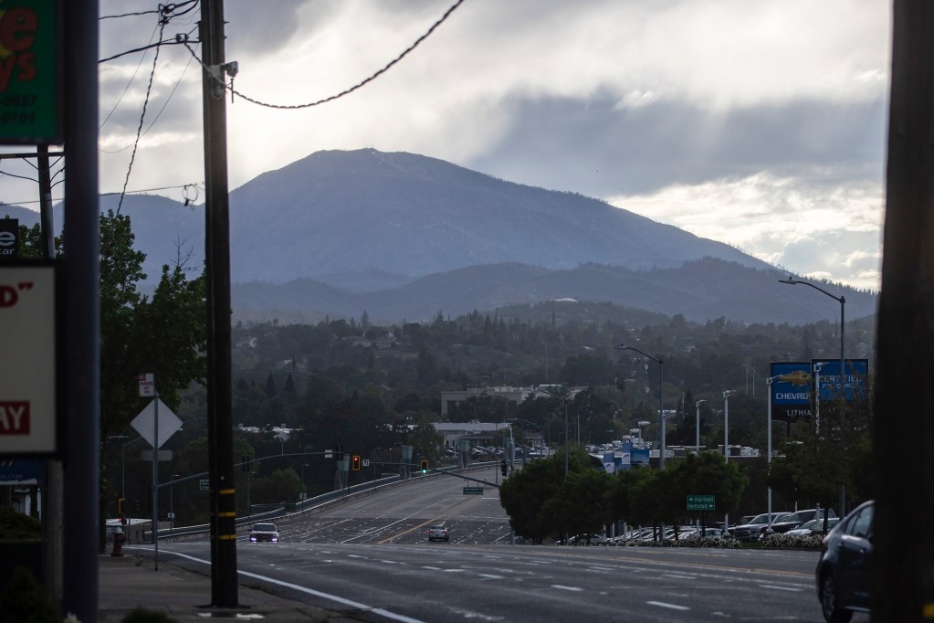 A view of a road leading into downtown Redding, with cars driving and a mountain range in the far background, while clouds create an overcast, giving a dark and gloomy scene.