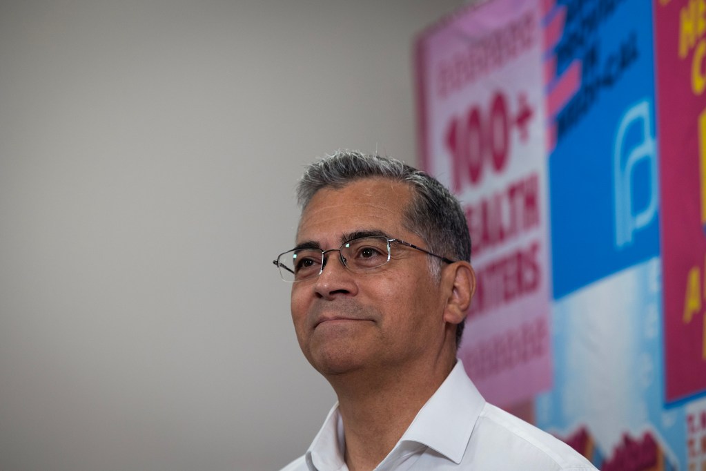 A man wearing glasses and a white collared shirt stands in front of colorful healthcare posters, looking slightly upward.
