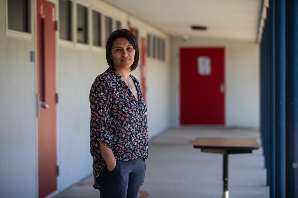 A person with short brown hair and wearing a black and pink flora blouse, stands in the walkway of an outdoor school building with red doors and blue columns.