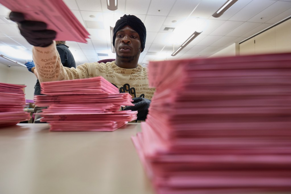 A worker wearing gloves sorts stacks of pink election ballots on a table. Piles of ballots fill the foreground and background, while the worker reaches across the table inside a brightly lit ballot-processing facility.