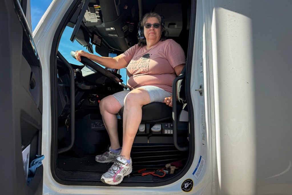 A long-haul truck driver sits in the open cab of a semi-truck, one hand resting on the steering wheel. Wearing sunglasses, a headset and casual clothing, she looks toward the camera from the driver's seat. Bright sunlight illuminates the truck doorway while the cab interior remains in shadow.