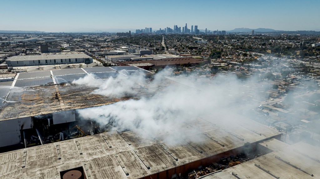 An aerial view shows smoke rising from a damaged warehouse roof as firefighters continue to battle a large industrial fire. The surrounding neighborhood stretches toward the downtown Los Angeles skyline in the distance, partially obscured by haze drifting across the scene.