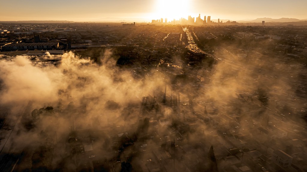An aerial view shows thick smoke drifting over an industrial neighborhood as the downtown Los Angeles skyline glows in the distance at sunset. The haze blankets homes, warehouses and streets below, with sunlight filtering through the smoke in golden beams.