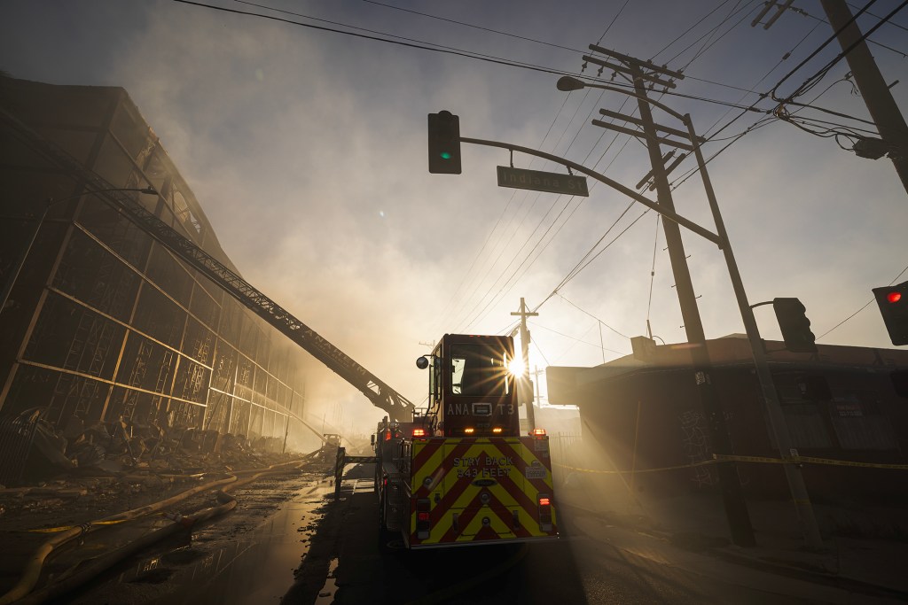 A ladder truck is parked beside a heavily damaged warehouse as firefighters continue battling a fire in the early morning. Smoke fills the air while sunlight breaks through the haze behind the truck, illuminating fire hoses, debris and the skeletal remains of the building.