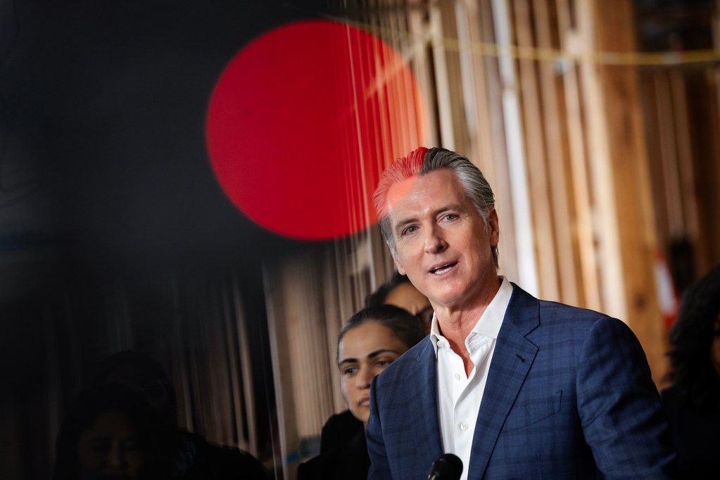 California Gov. Gavin Newsom wears a blue suit jacket and white shirt while speaking indoors, with several people standing behind him and a large red circular light in the foreground.