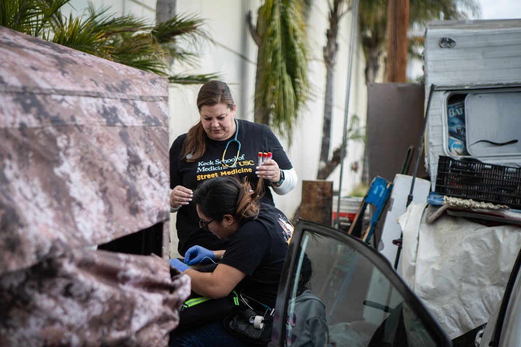 Two people provide medical care beside a parked vehicle while another person stands nearby holding medical supplies.