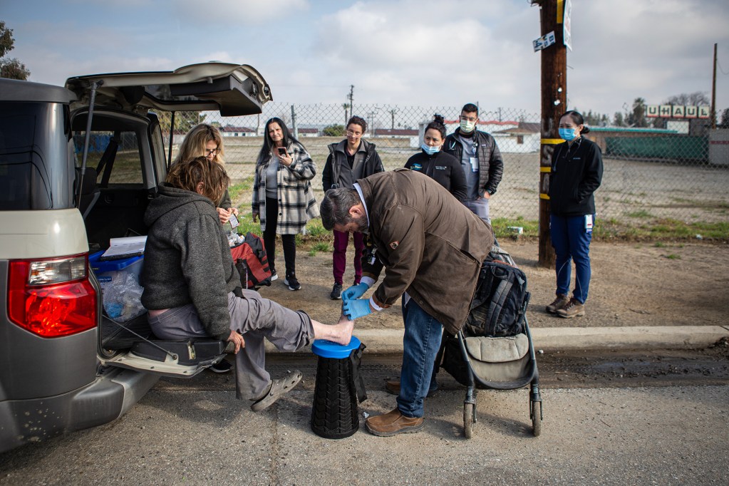 A healthcare worker treats a person's foot while they sit on the open tailgate of an SUV as several people watch nearby.