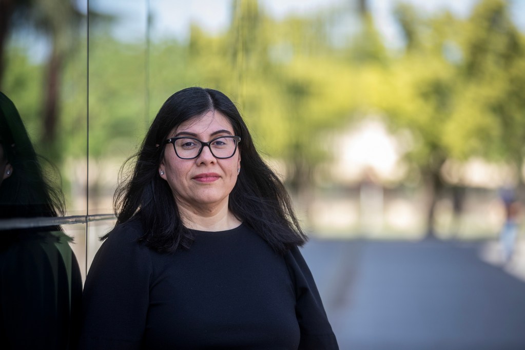 A woman with shoulder-length dark hair and glasses stands against a reflective glass wall, looking toward the camera, with trees and a walkway softly blurred in the background.