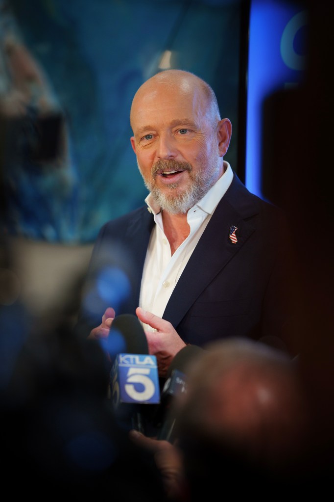Steve Hilton speaks with reporters during a campaign event, smiling as television news microphones are held toward him while photographers and audience members frame the foreground.