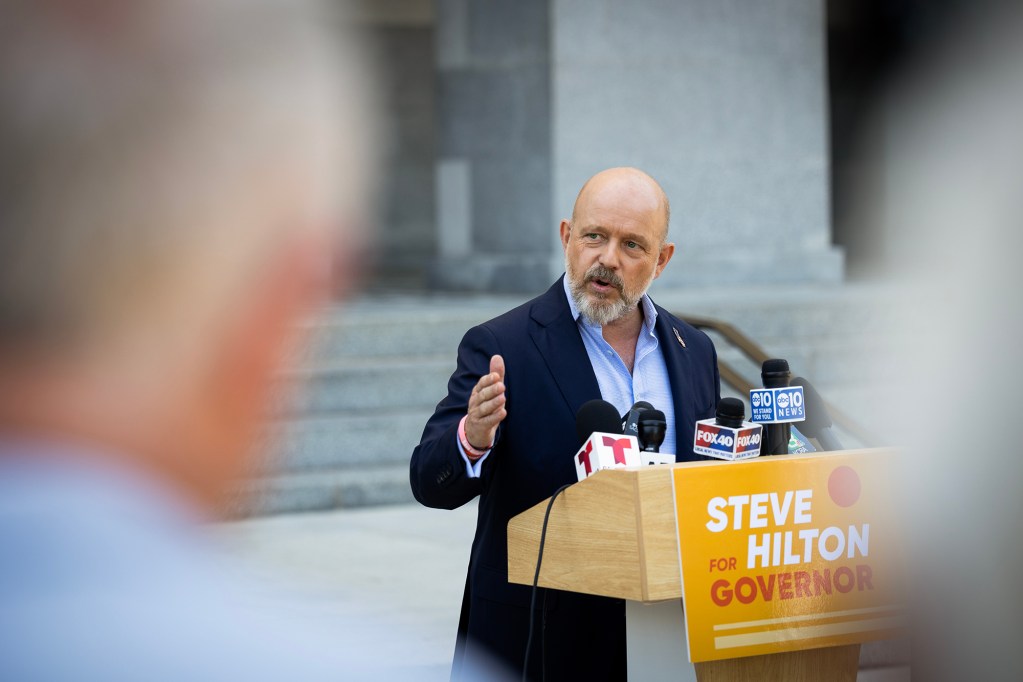 Steve Hilton speaks at a podium during a news conference outside the California State Capitol, gesturing as he addresses reporters with several television news microphones mounted on the lectern.