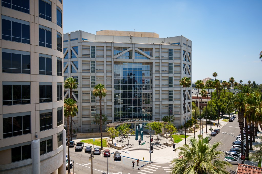 A beige and blue glass mirror building on the corner of a street lined with palm trees.