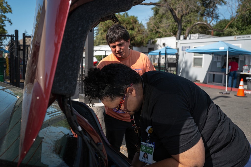 Two people stand at the open trunk of a vehicle, looking through supplies in a parking lot.
