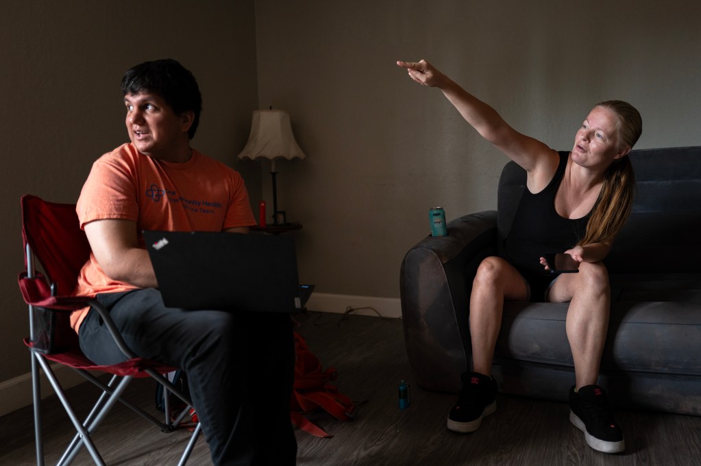 A woman sits on a couch pointing across the room while speaking with a member of a street medicine team, who listens with a laptop open on their lap inside her apartment.
