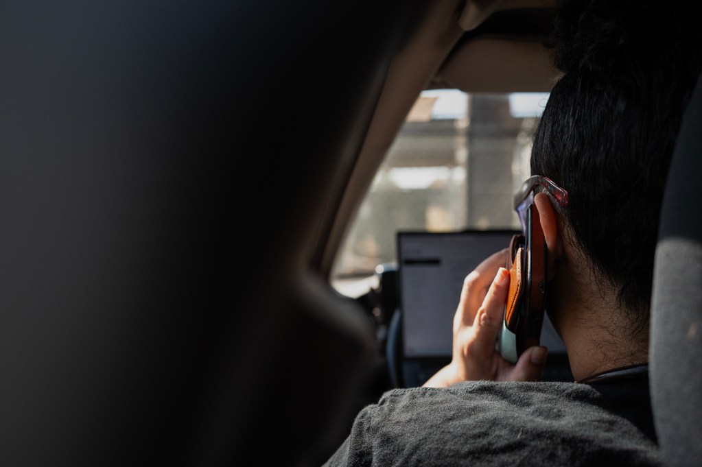 A person talks on a cellphone while sitting in the front seat of a vehicle, with a laptop open on the dashboard.