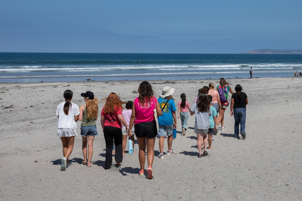 A view from being of middle school-aged children and adult camp counselors walking along the shore and towards a beach.