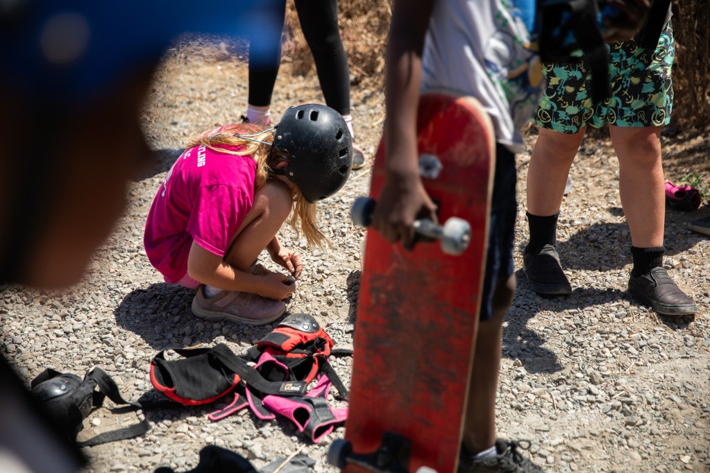A middle school-aged child wearing a skateboard helmet and a pink shirt, squats down to the gravel as they hold a rock in their hands. Other children can be seen in the foreground, alongside equipment such as skateboards and knee pads.