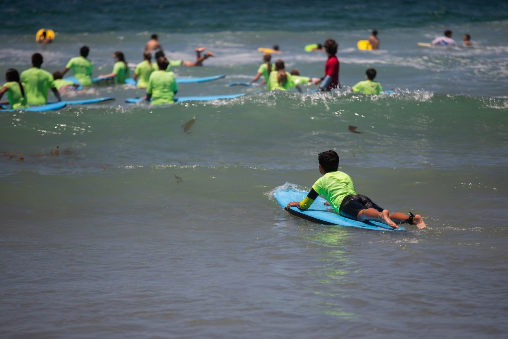 A middle school-aged child wearing a neon green shirt, lays on a boogie board while riding a small wave. Other chidlren can be seen in the background holding boogie boards.