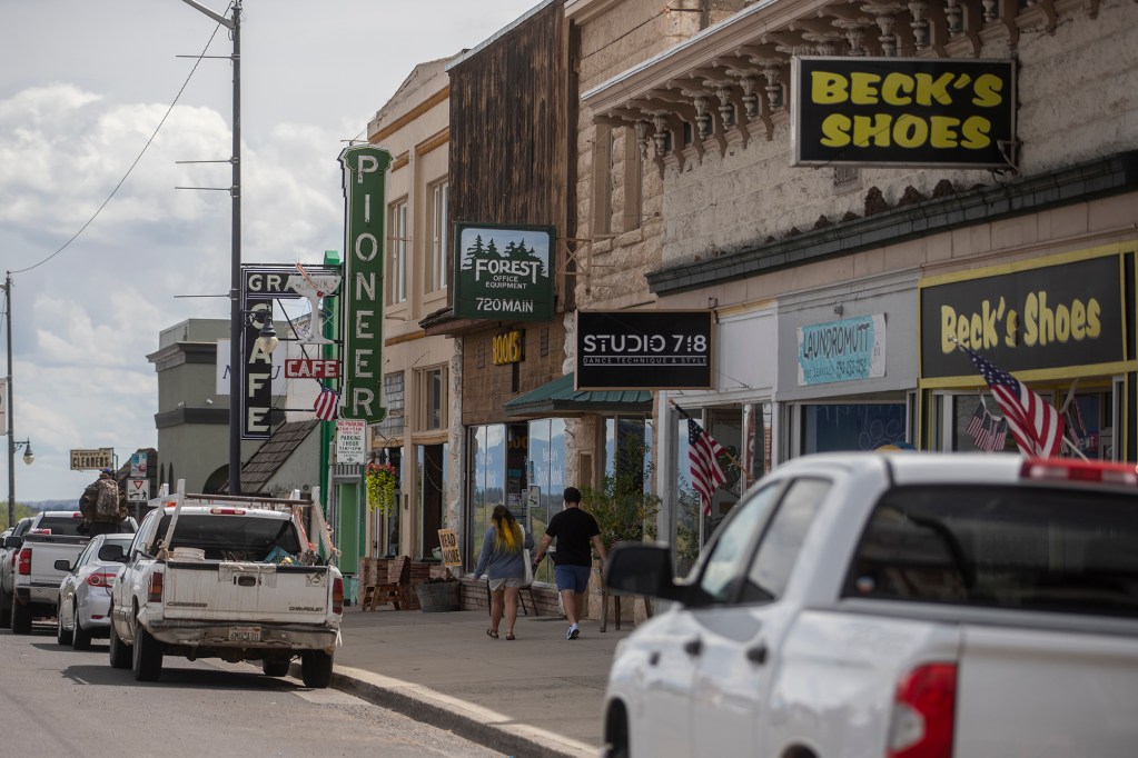A couple of people walk down the street along the sidewalk in front of a row of small businesses with their signs hanging from the buildings.