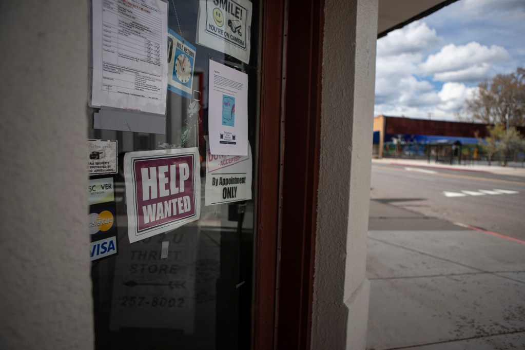 A help-wanted sign hangs with other flyers and signs in the window of a glass door of a small business, with the main road through a small town in the background.