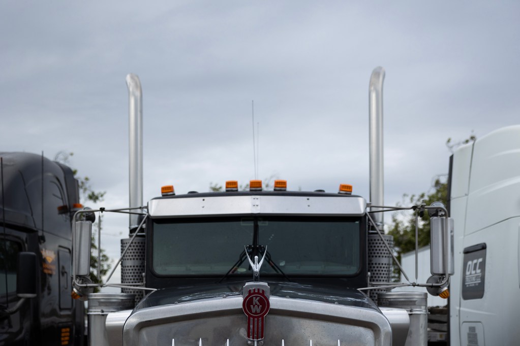 A view of the front of a semi truck from the upper cab with two silver exhaust pipes going up with a grey overcast sky in the background,
