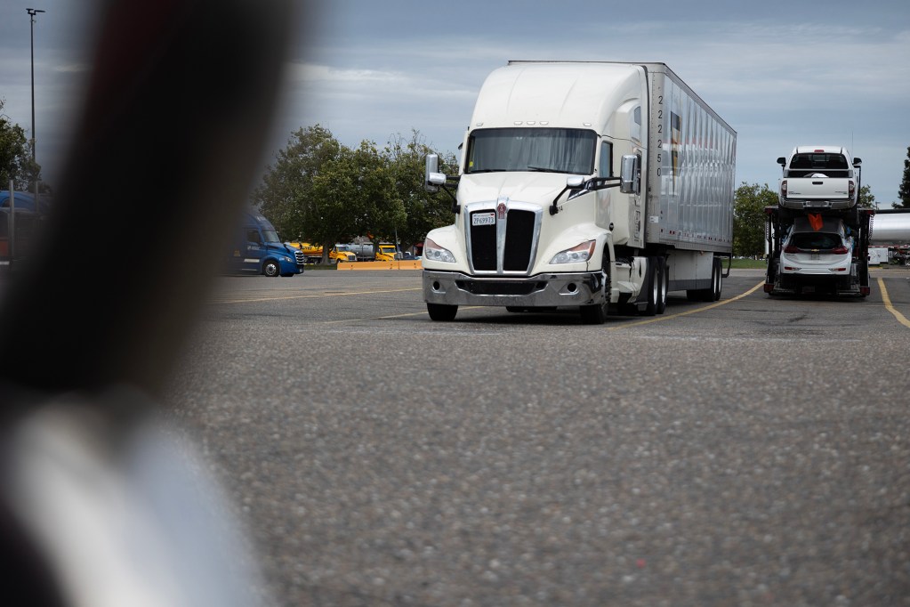 A white semi-truck parked in a parking lot with other semi-trucks and a truck hauling vehicles around.