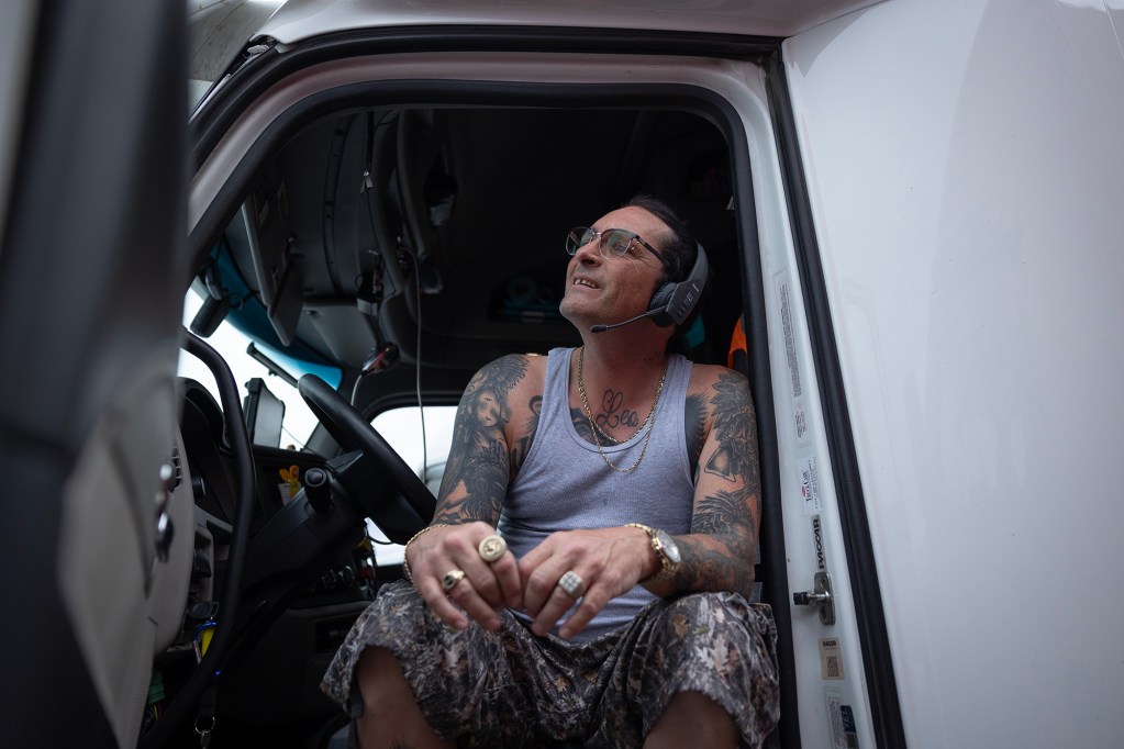 A person wearing glasses and a headset sits in the driver's seat of a semi-truck with the door open. The person has a series of tattoos visible on his upper body and wears an assortment of rings on both hands.