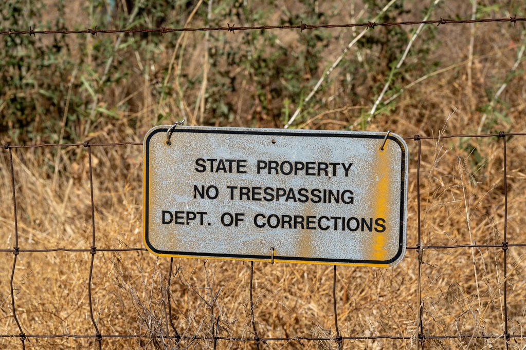 A close-up shot of a worn-out sign reading "State Property No Trespassing Dept. of Corrections," pinned to a barbed wire fence with dried brush in the background.